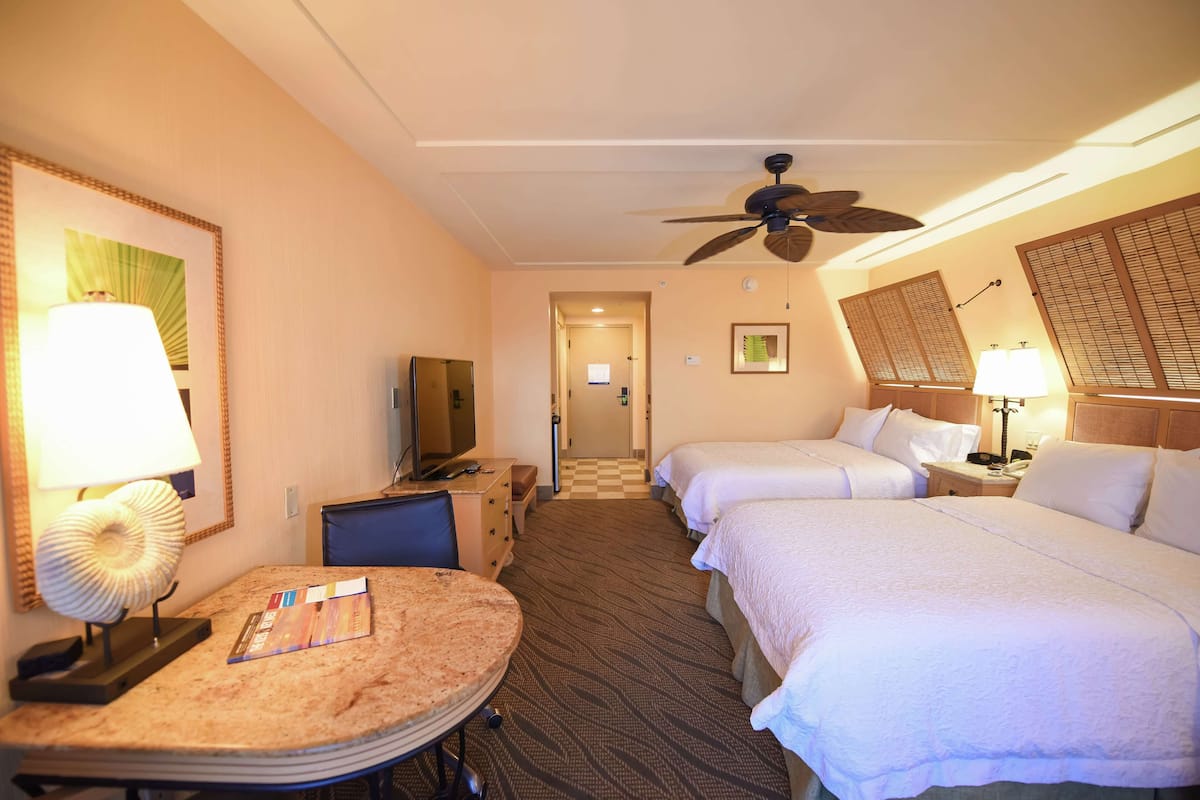 A hotel room with two beds, a desk with a lamp, a television, and a ceiling fan.