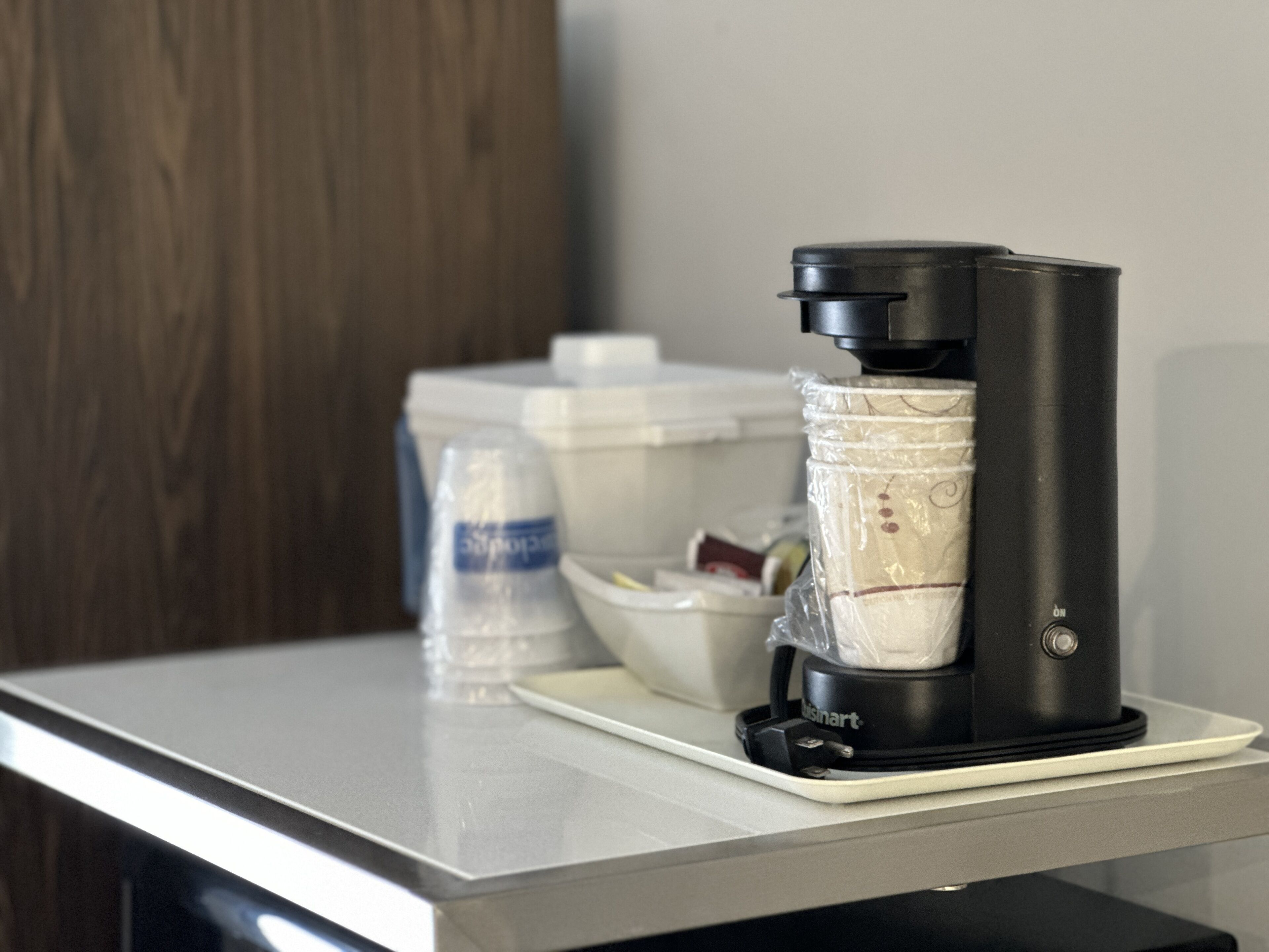 Una cafetera sobre una bandeja blanca, un dispensador de agua y un recipiente blanco con tapa en una habitación de hotel.
