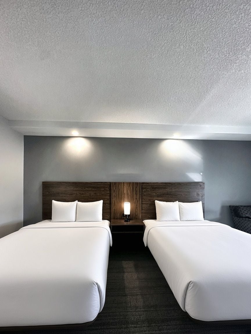 A hotel room with two beds, a wooden headboard, a nightstand with a lamp, and a wall-mounted light fixture.