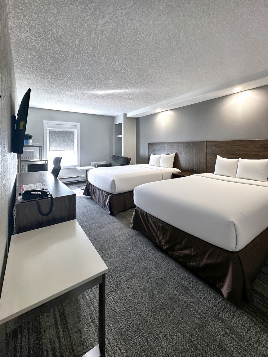 A hotel room with two beds, a desk, a chair, a TV, and a window.