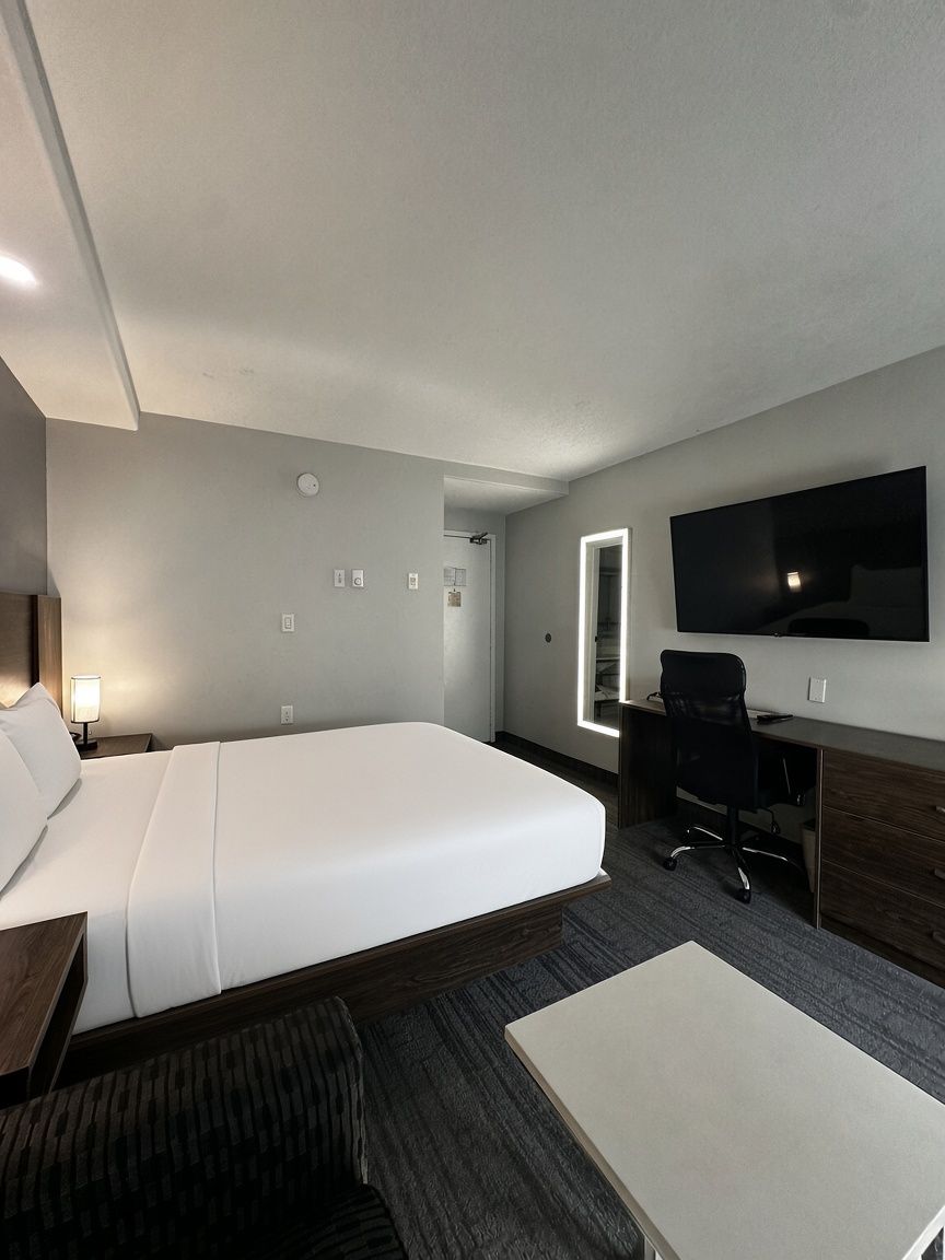A hotel room with a large bed, a desk, a chair, a flat-screen TV, and a bedside lamp.