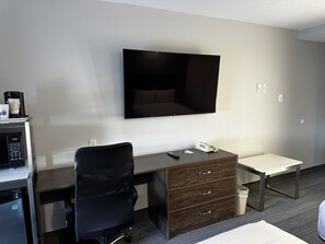 A hotel room with a flat-screen TV mounted on the wall, a desk with a chair, a small table, and a trash bin. - Travelodge by Wyndham Regina (Regina)