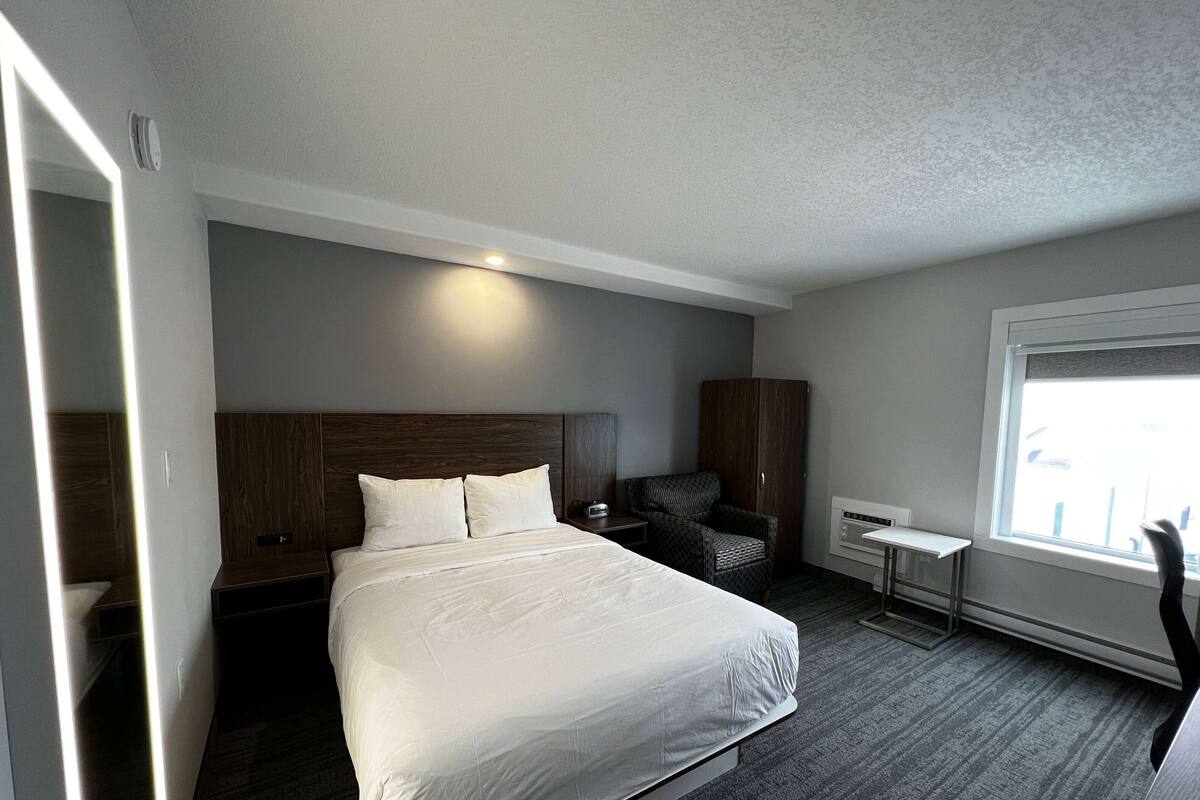 Una habitación de hotel moderna con una cama grande, un escritorio pequeño, una silla, un armario y una ventana con luz natural.