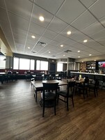 Un restaurante con varias mesas y sillas, una zona de bar y una pantalla de televisión mostrando un evento deportivo.