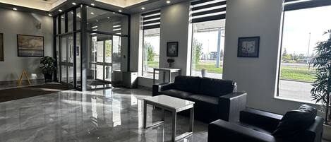 A spacious lobby with a modern design, featuring a marble floor, black leather seating, and large windows offering views of the outside.