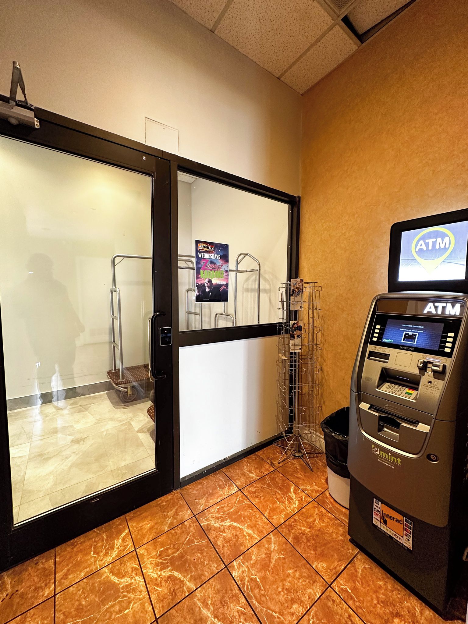 An ATM machine with a screen, a glass door entrance, and a poster advertising a karaoke event.