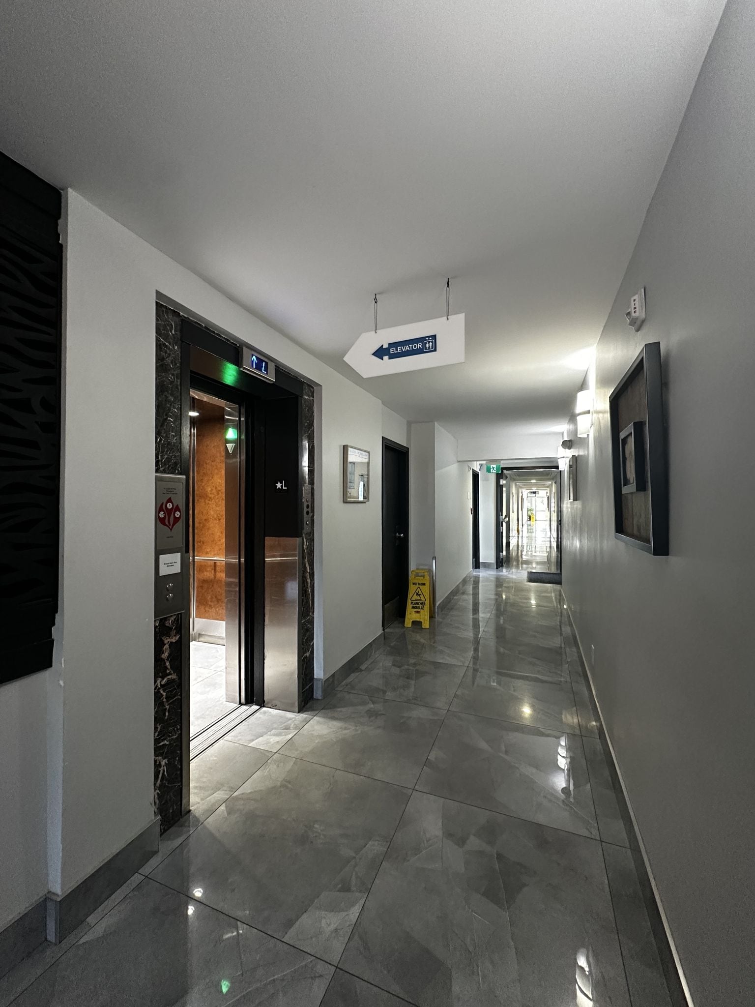 A hotel corridor with an elevator, directional signs, and a wet floor sign.