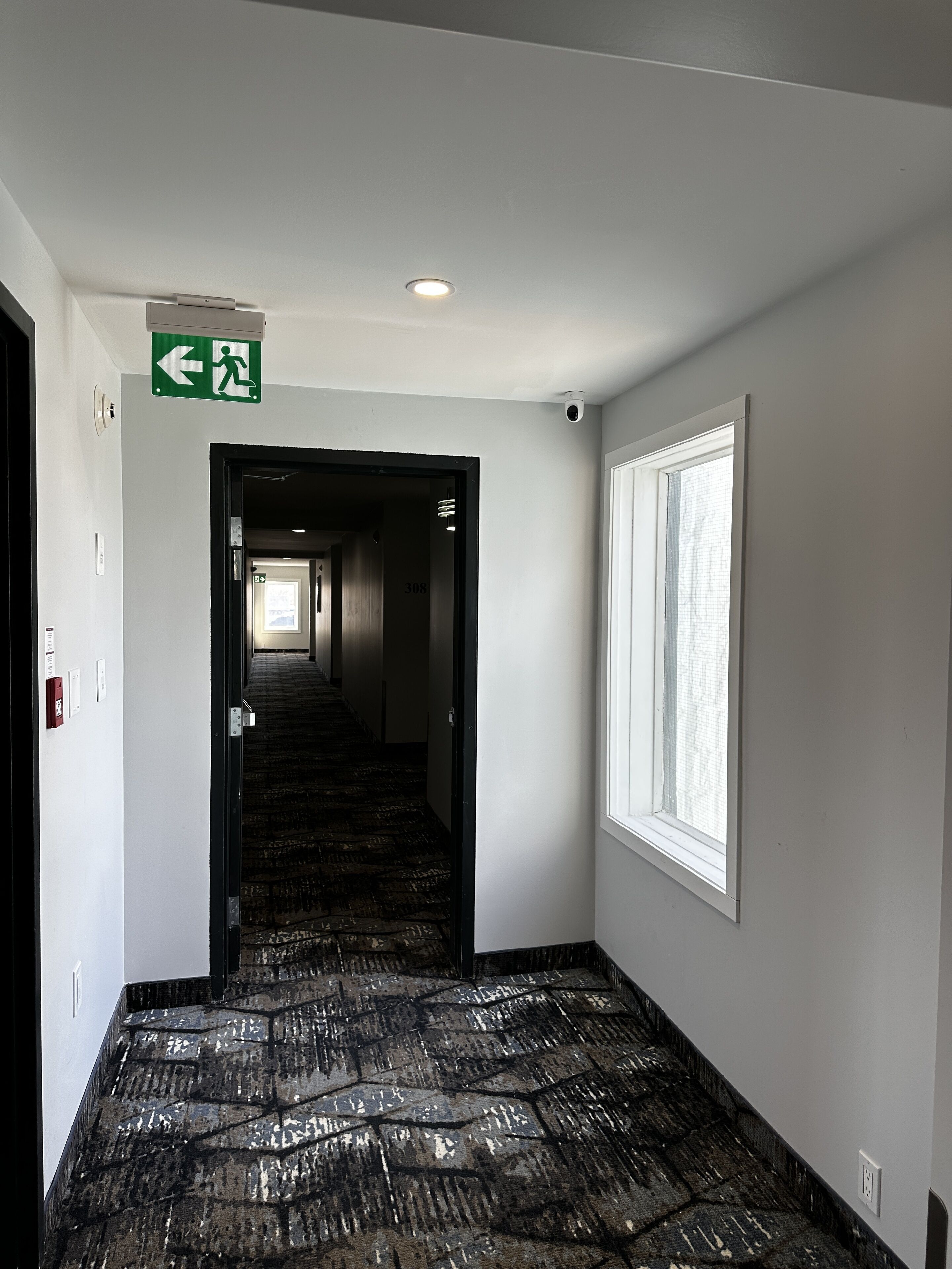 A hotel hallway with a carpeted floor, a window on the right, and an exit sign on the left.