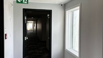A hotel hallway with a carpeted floor, a window on the right, and an exit sign on the left.