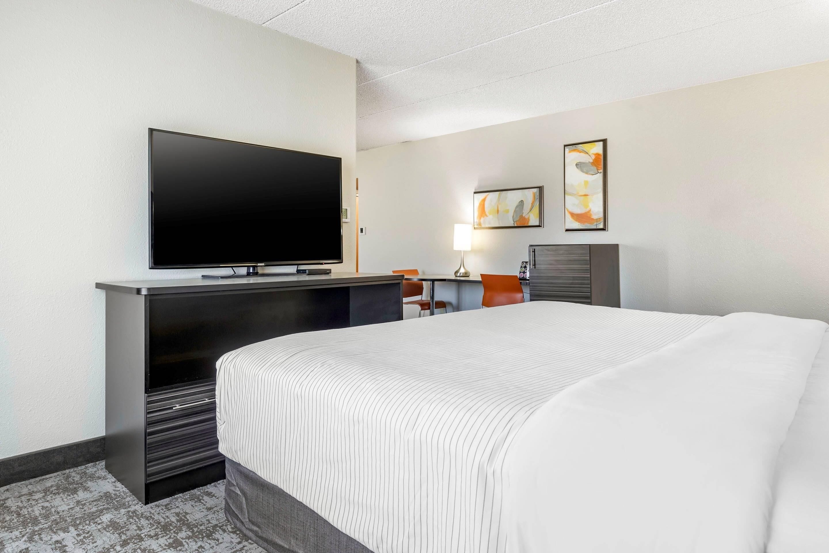 A hotel room with a large bed, a flat-screen TV mounted on a cabinet, a desk with a chair, and two framed artworks on the wall.