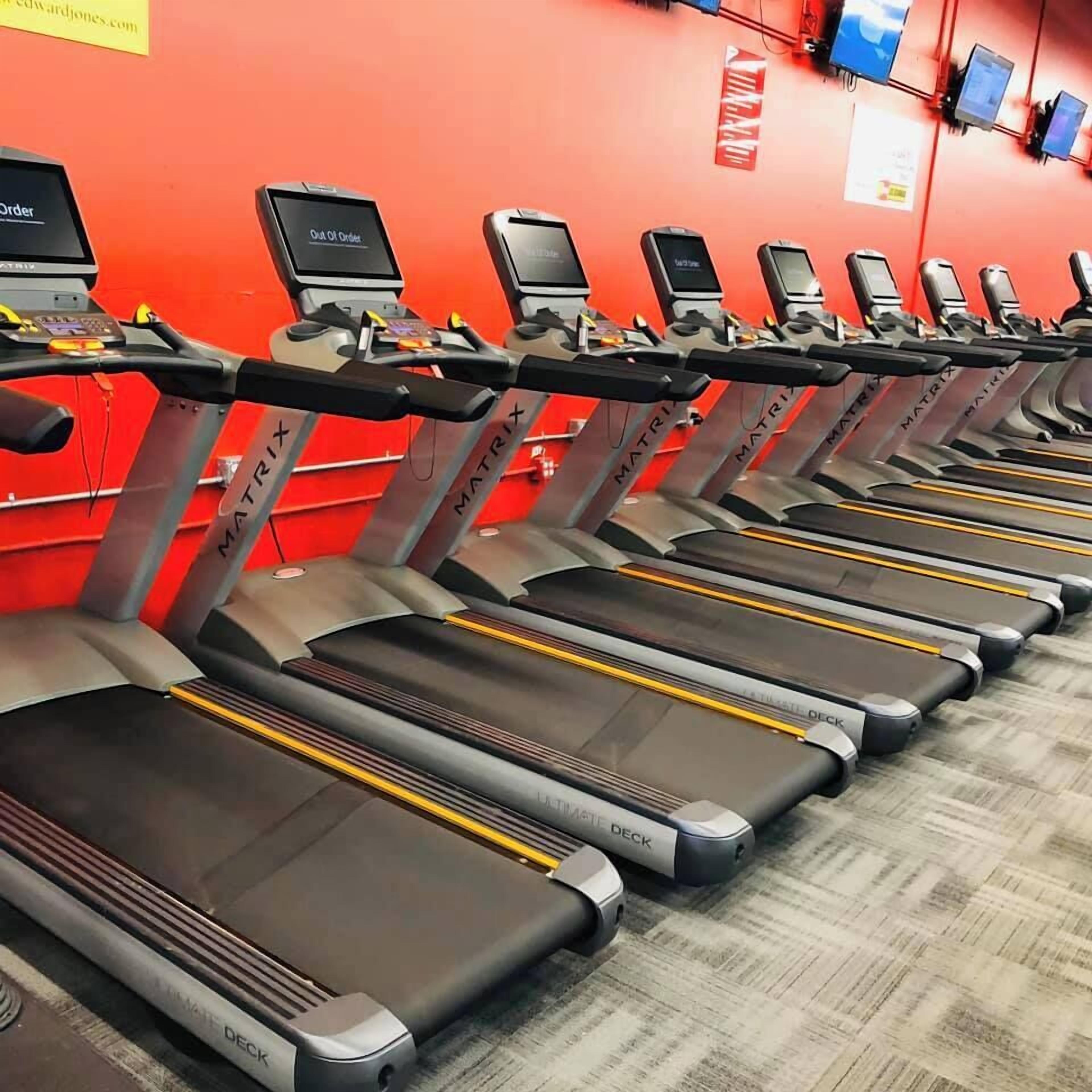 Une rangée de tapis de course dans une salle de sport, avec un mur rouge et un sol recouvert de moquette.