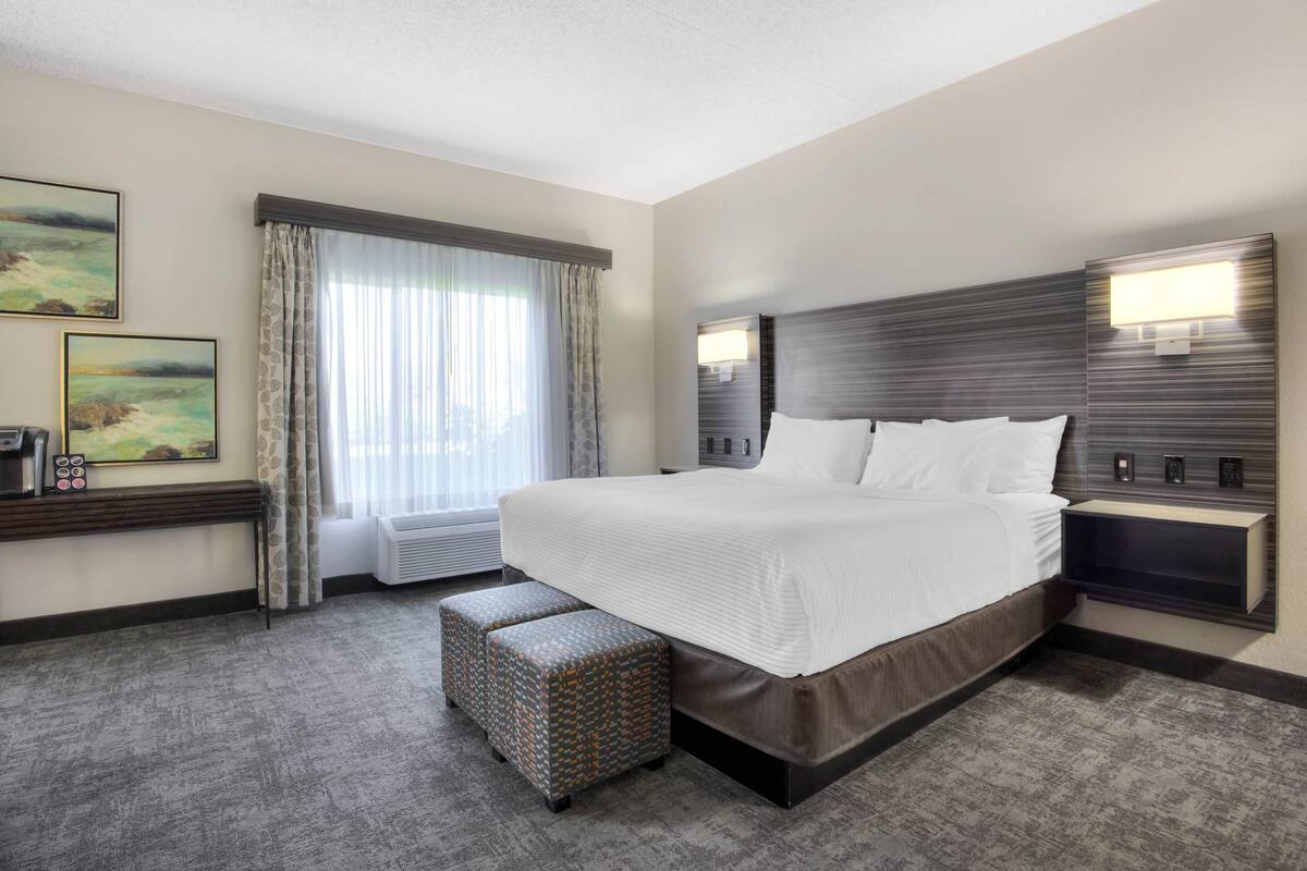 A modern hotel room with a large bed, a bedside table, a wall-mounted lamp, and a window with curtains.