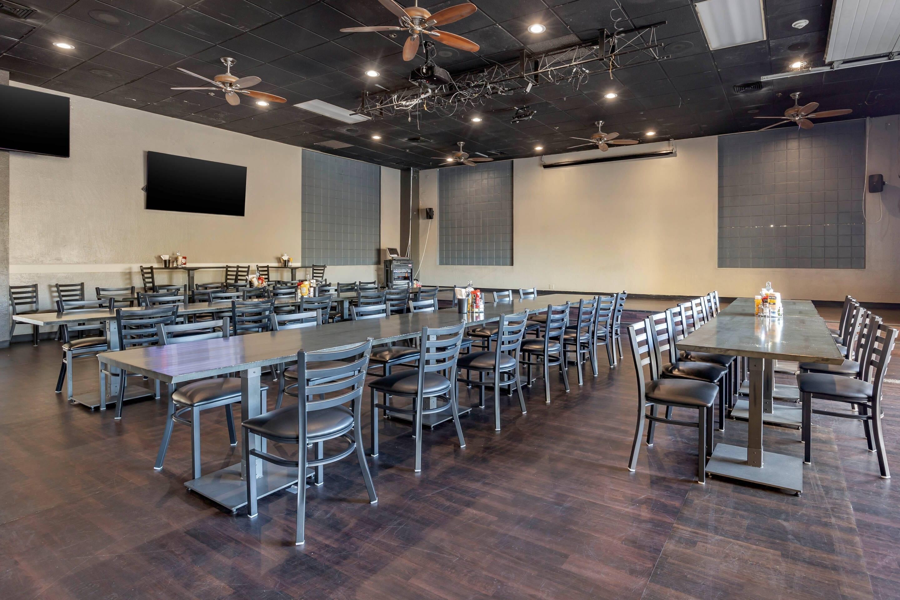 A large conference room with multiple tables and chairs, ceiling fans, and a flat-screen TV mounted on the wall.