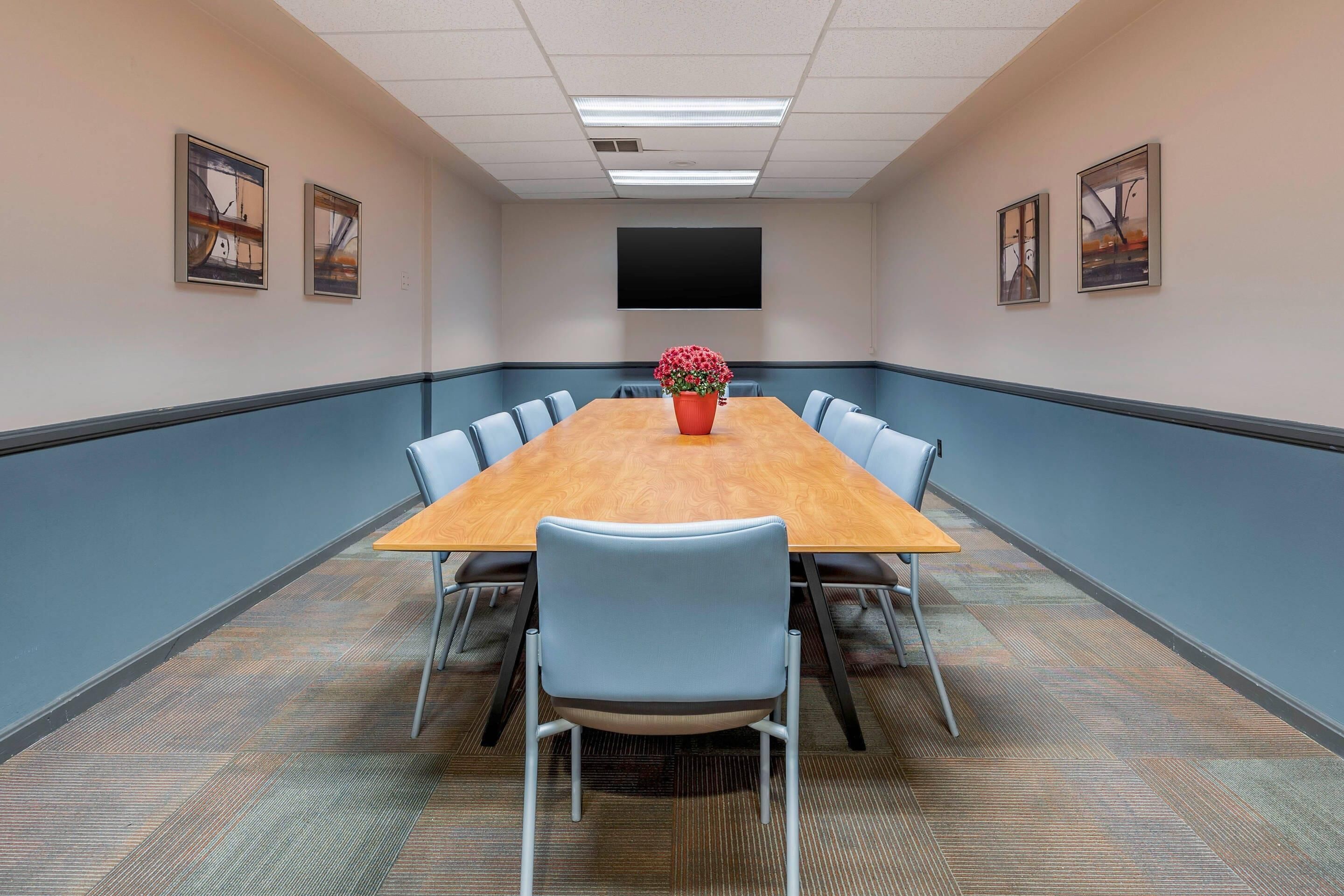 A conference room with a long table, blue chairs, a red flower vase, and framed artwork on the walls.