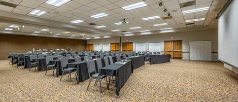 A conference room with rows of tables and chairs, carpeted floor, ceiling lights, and a projector.