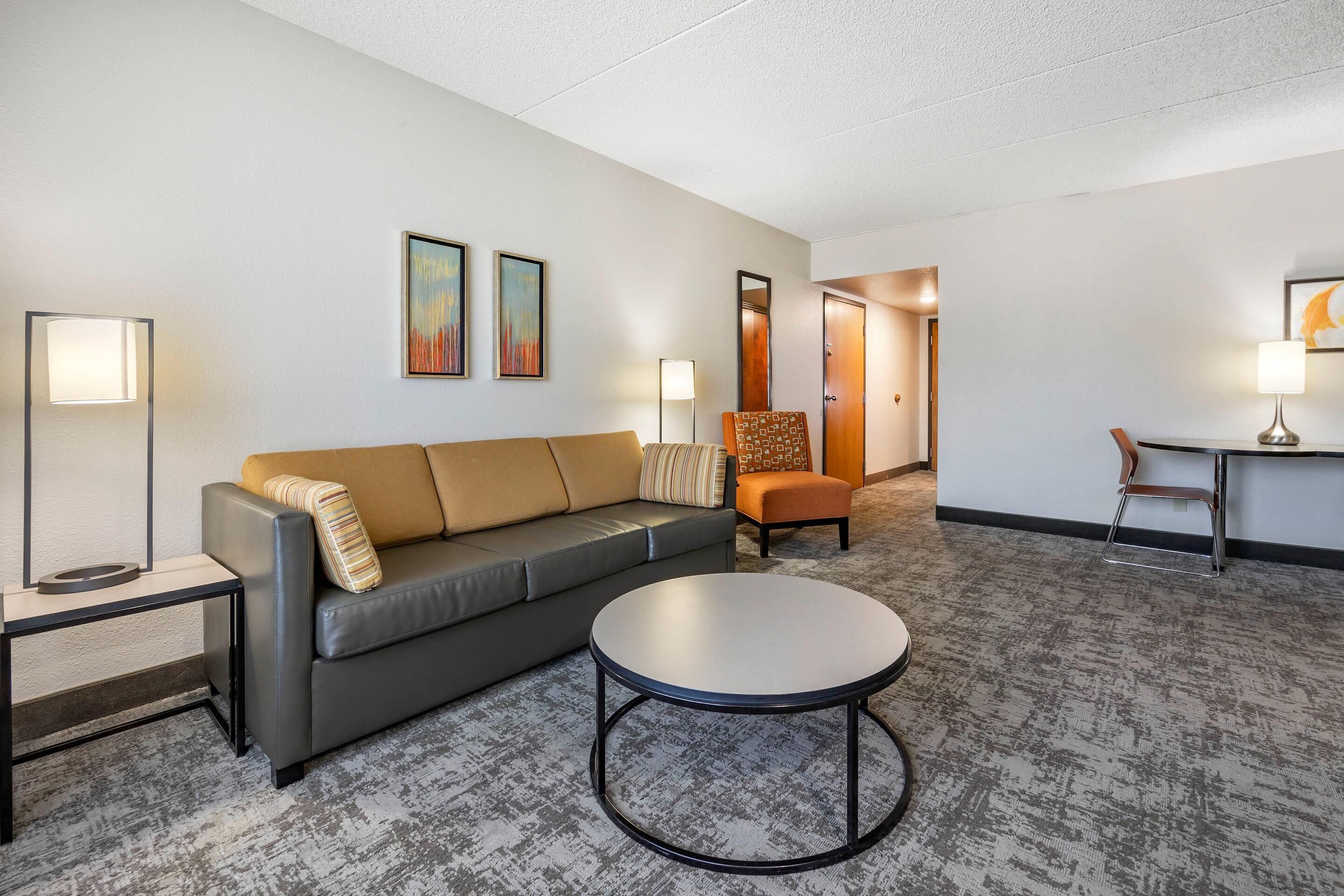 A hotel room with a gray sofa, a round coffee table, a side table, a chair, and two wall-mounted lamps.