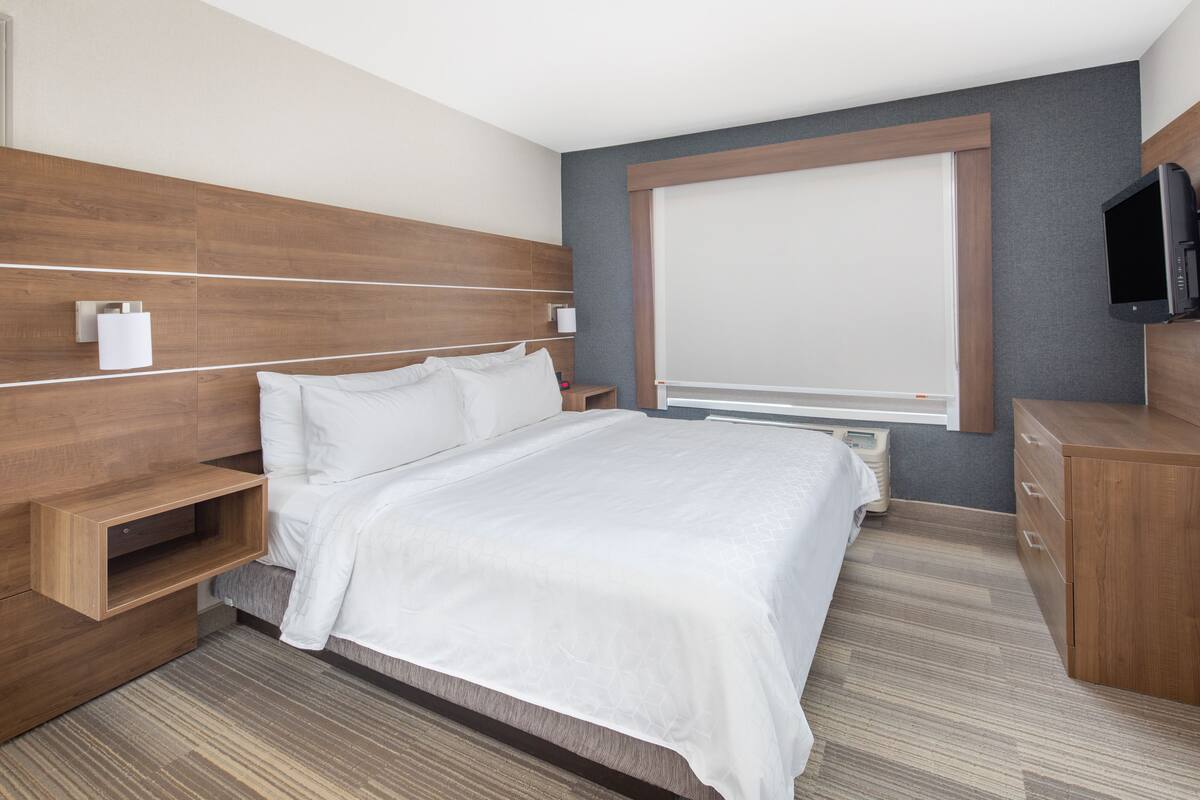 Une chambre d’hôtel moderne avec une tête de lit en bois, un grand lit avec du linge de maison blanc, un téléviseur à écran plat et un radiateur intégré.
