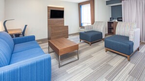 Une chambre d’hôtel moderne avec un canapé bleu, une table basse en bois, deux fauteuils à motifs, un téléviseur fixé à un meuble en bois et un petit coin cuisine.