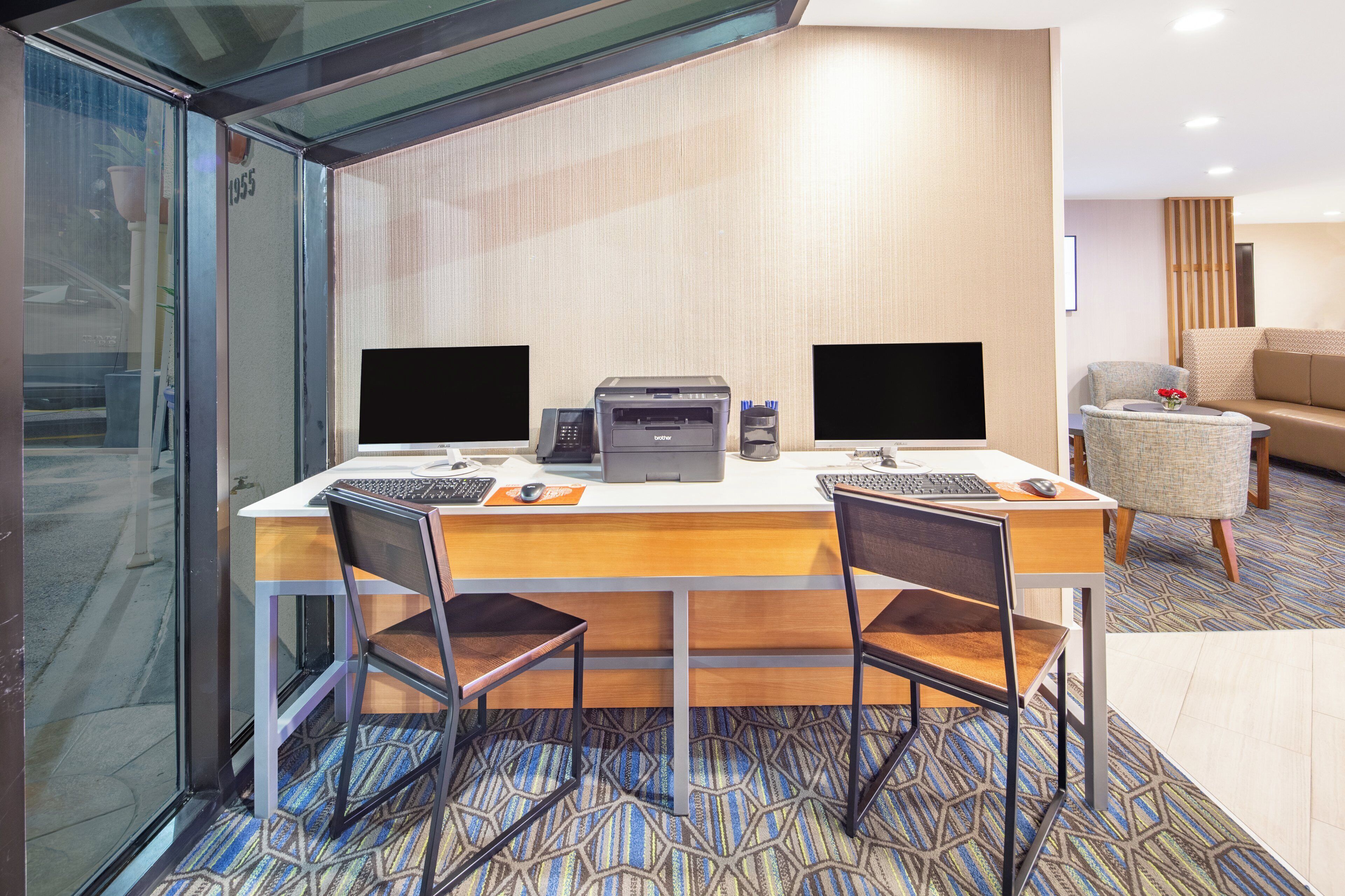 A hotel room with a desk, two monitors, a printer, and a chair.