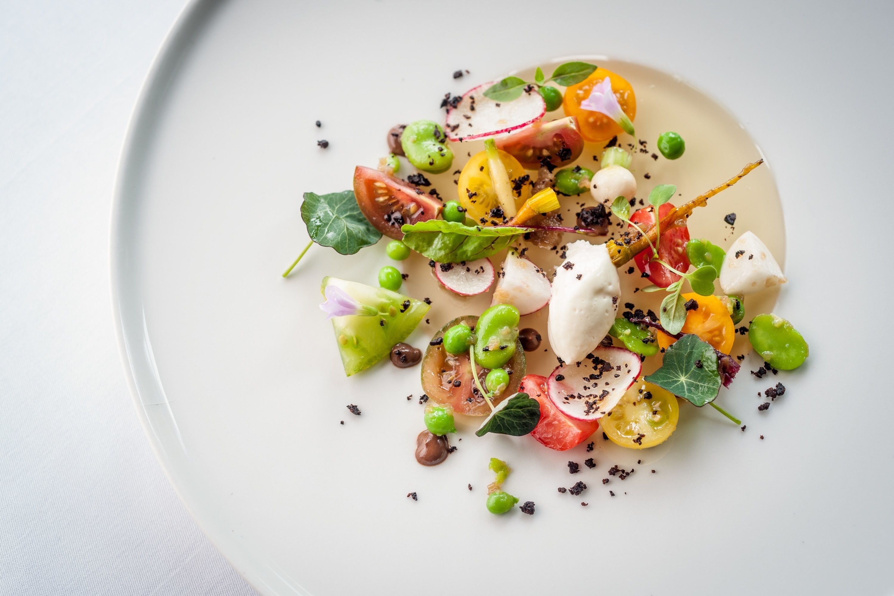 Un plato blanco con una ensalada colorida, que incluye tomates, guisantes y rábanos, adornada con semillas negras y una hoja verde.