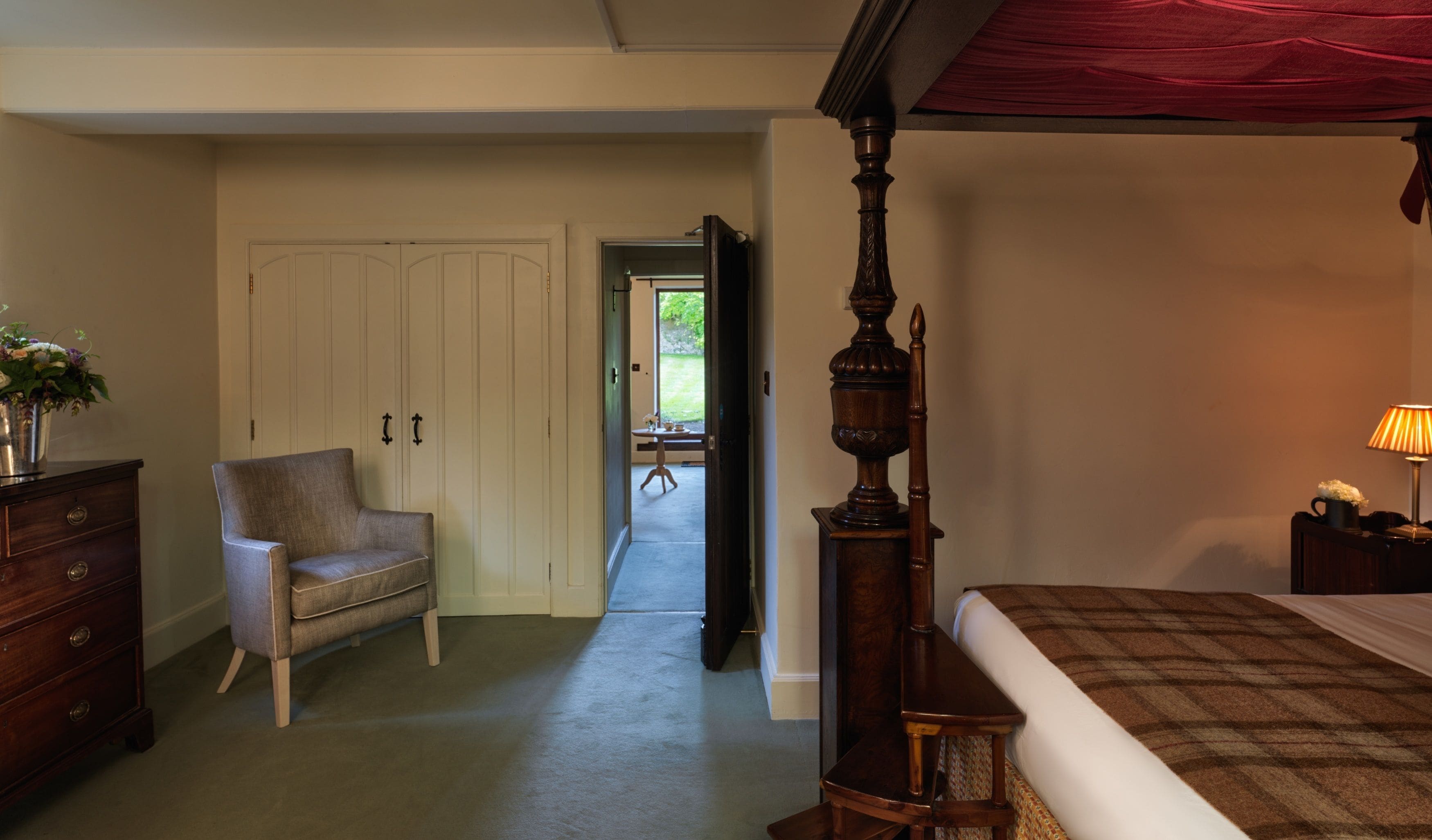 A bedroom with a four-poster bed, a chair, a dresser, and a nightstand with a lamp.