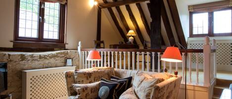 A cozy room with a high ceiling, exposed beams, a stone wall, a cushioned sofa, a wooden table, and two lamps.
