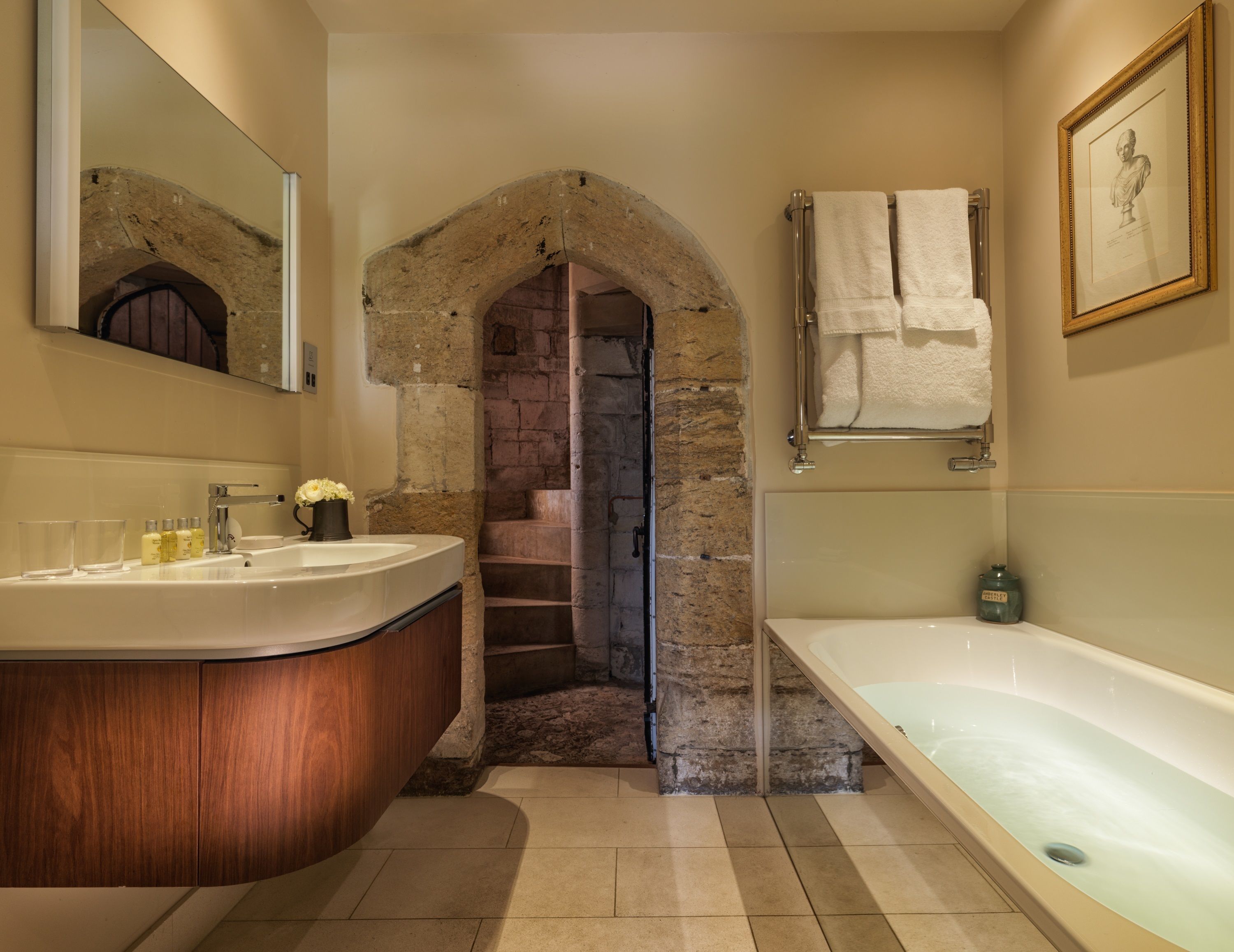 A bathroom with a stone archway leading to a staircase, a wooden vanity, a bathtub, and a framed portrait on the wall.