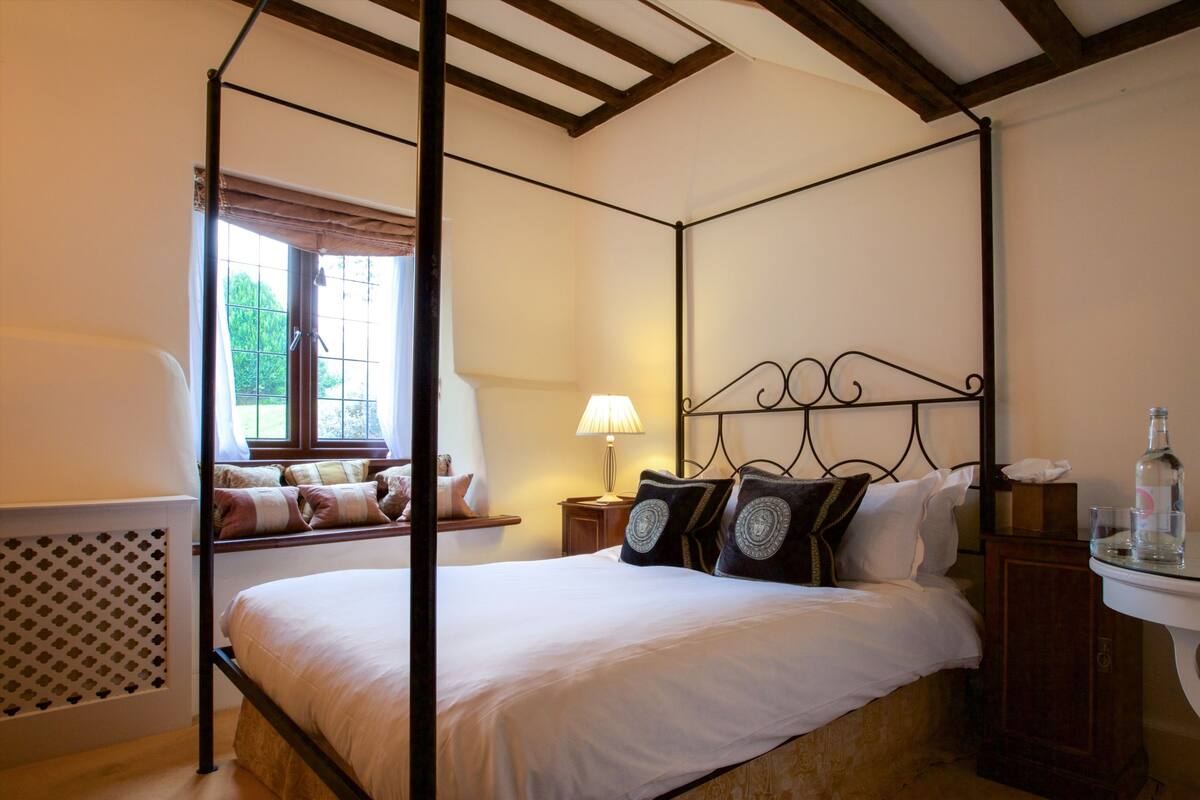 A four-poster bed with a canopy, a bedside table with a lamp, and a window with a view of greenery.