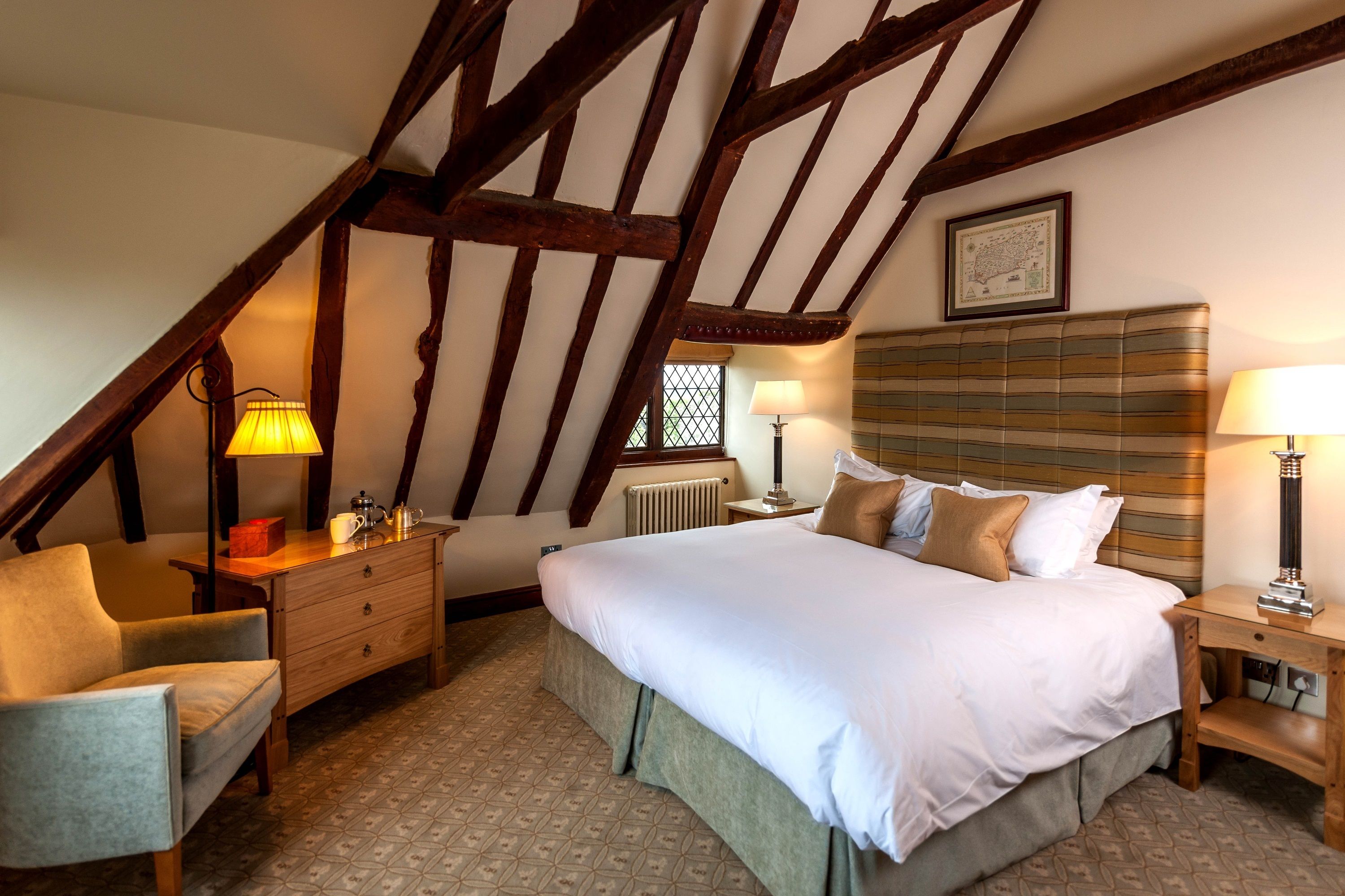 A bedroom with a sloped ceiling, a bed with white bedding, a wooden dresser, a chair, and two bedside lamps.