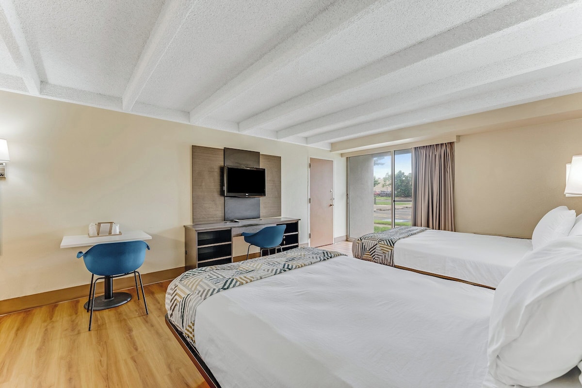 A hotel room with two beds, a desk with a chair, a TV, and a large window with curtains.