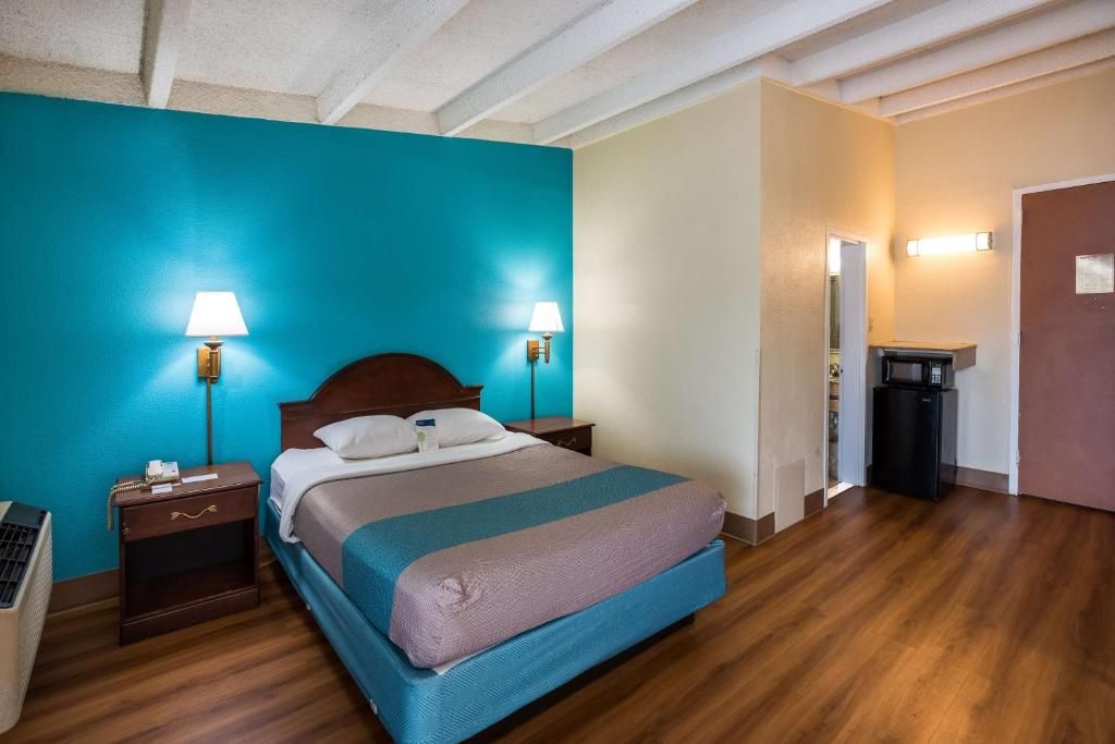 A hotel room with a bed, bedside lamps, a nightstand, a trash bin, and a door leading to another room.