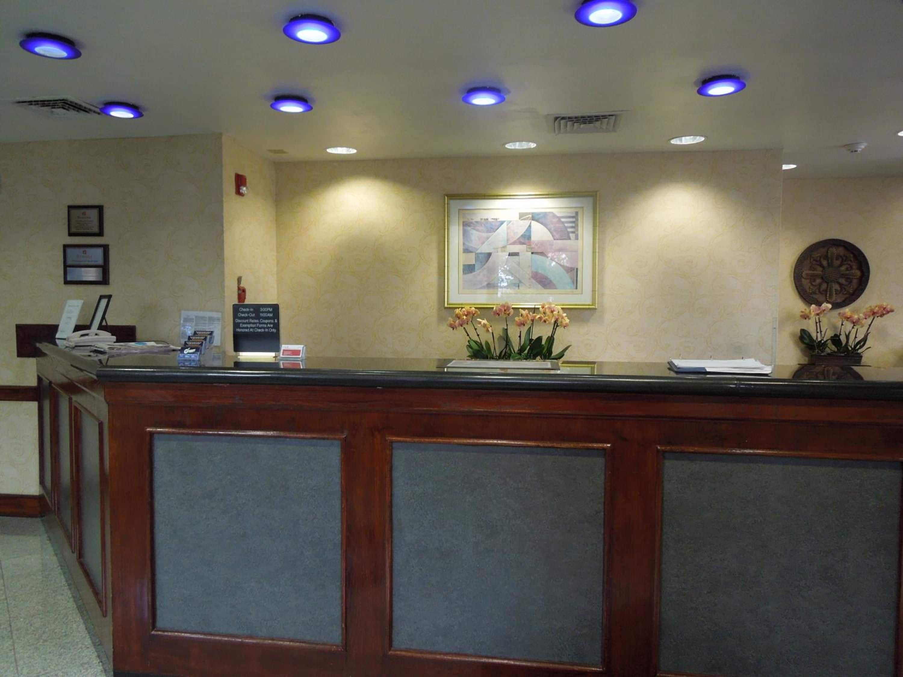 Un mostrador de recepción de hotel con una obra de arte enmarcada encima, un reloj y un jarrón con flores.