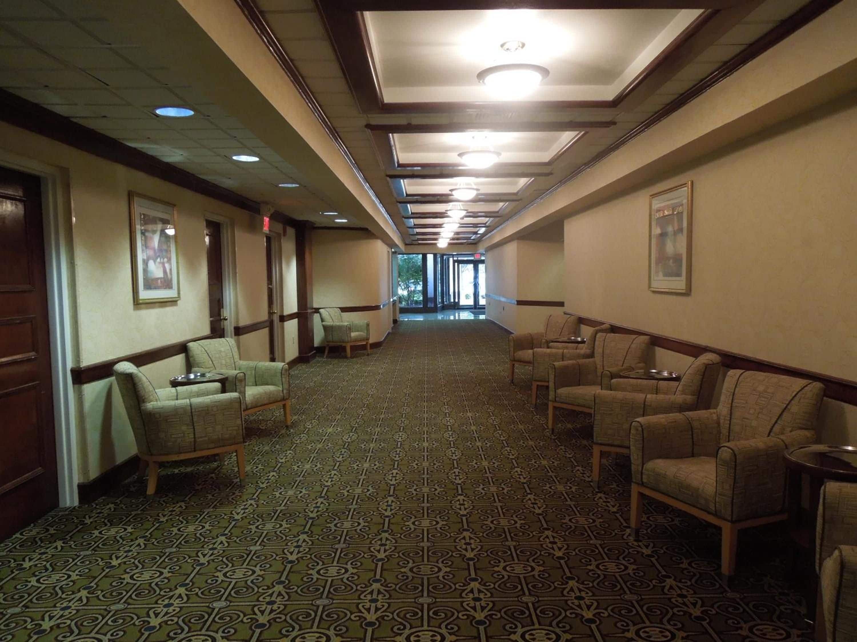 A hotel corridor with patterned carpet, chairs, and framed pictures on the walls.