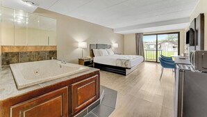 A modern hotel room with a large bathtub, a bed with a headboard, a desk with a chair, and a balcony with a view.