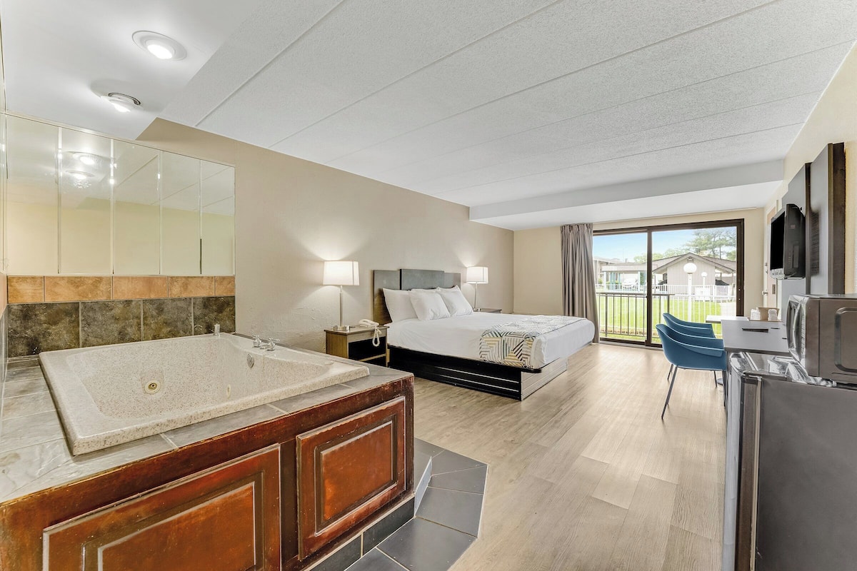 A modern hotel room with a large bathtub, a bed with a headboard, a desk with a chair, and a balcony with a view.