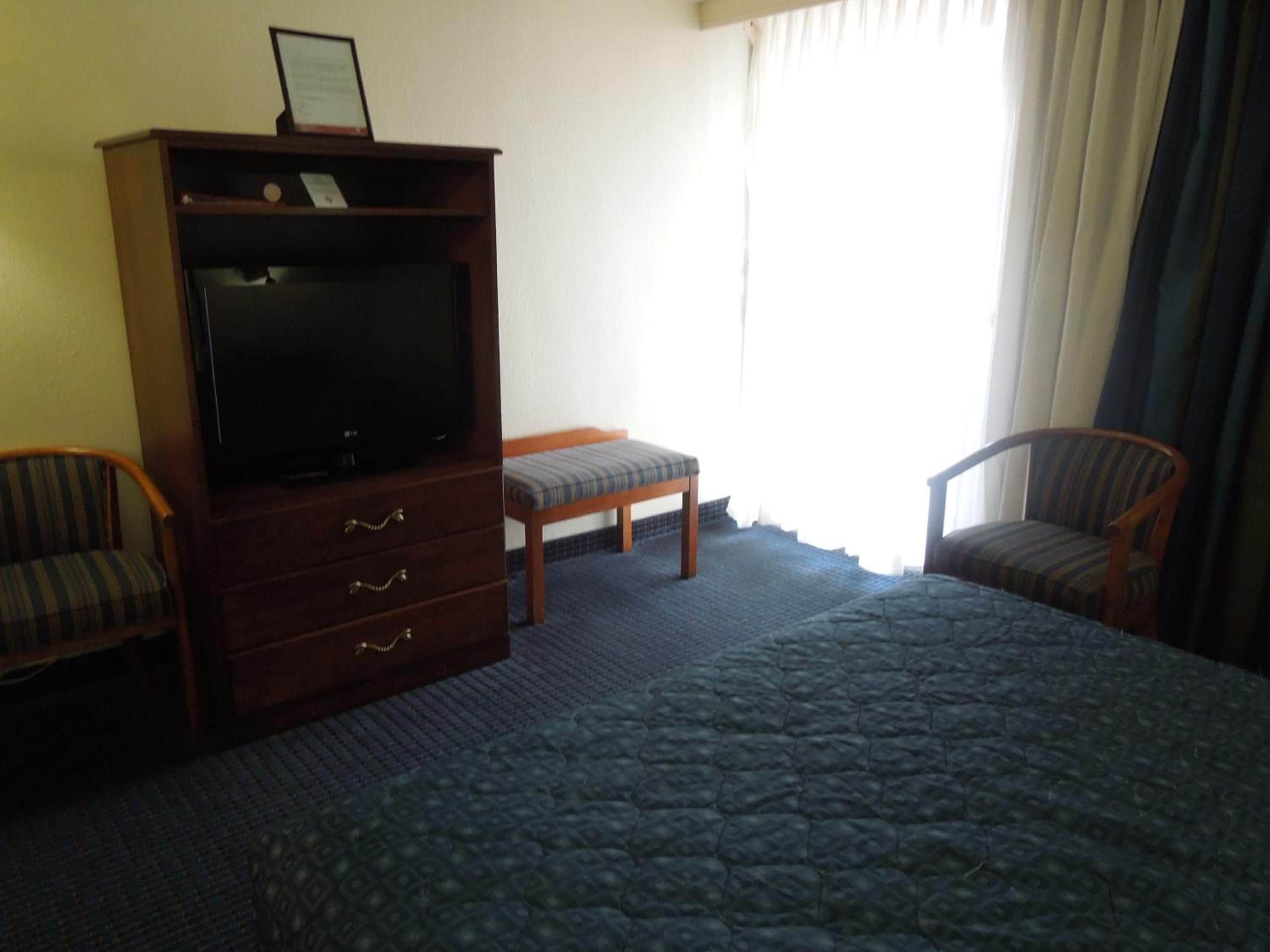 A hotel room with a bed, a television on a wooden cabinet, two upholstered chairs, and a window with curtains.
