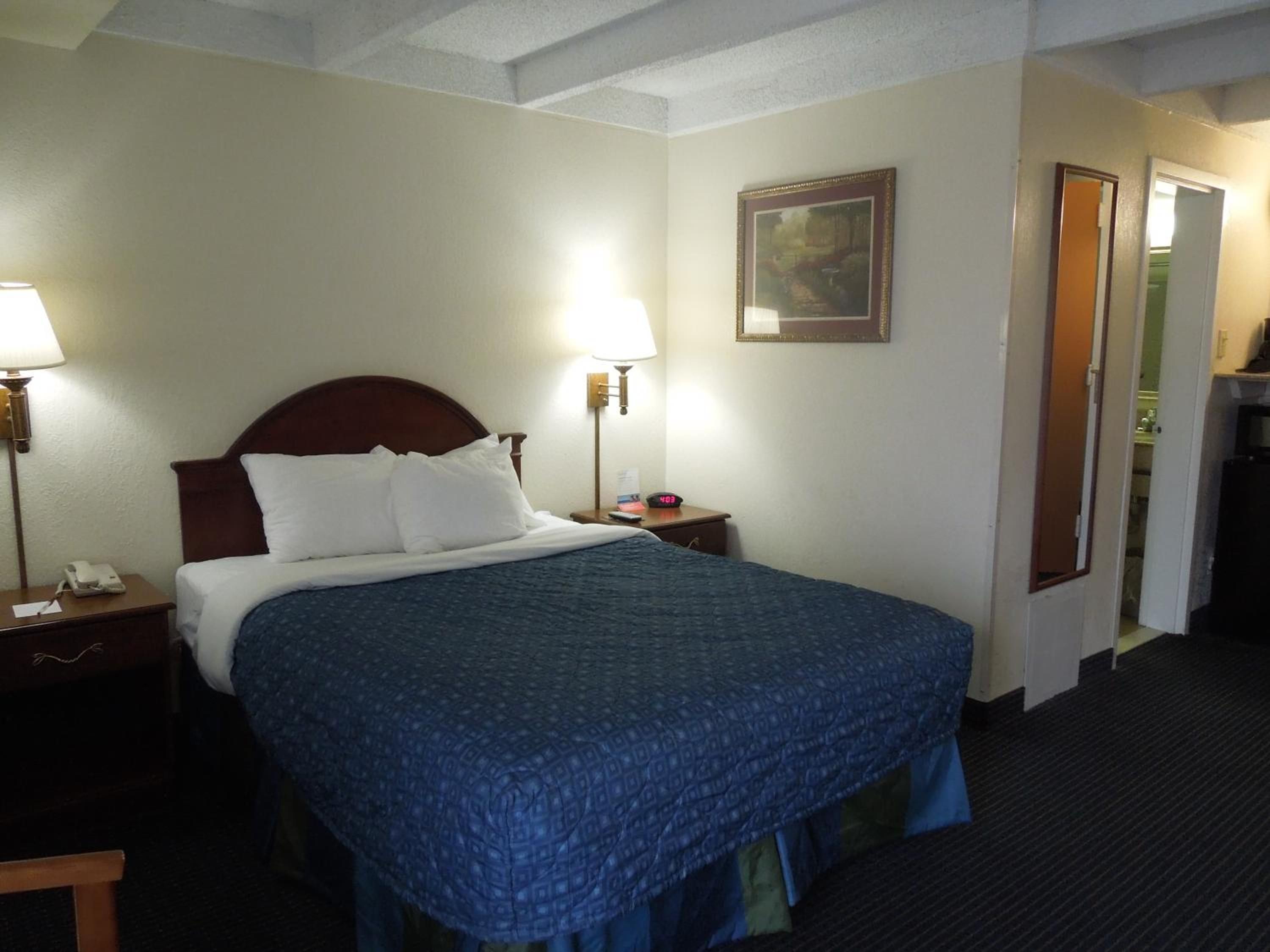 Habitación de hotel con cama, lámparas de noche, mesita de noche, un reloj y una puerta que da a un baño.
