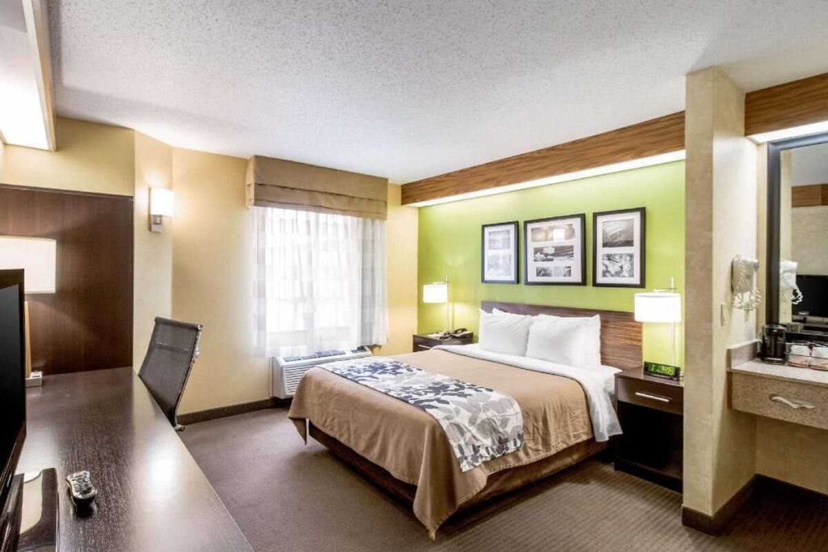 Una habitación de hotel con una cama, mesitas de noche, un escritorio con una silla, un televisor y un ventanal con cortinas.