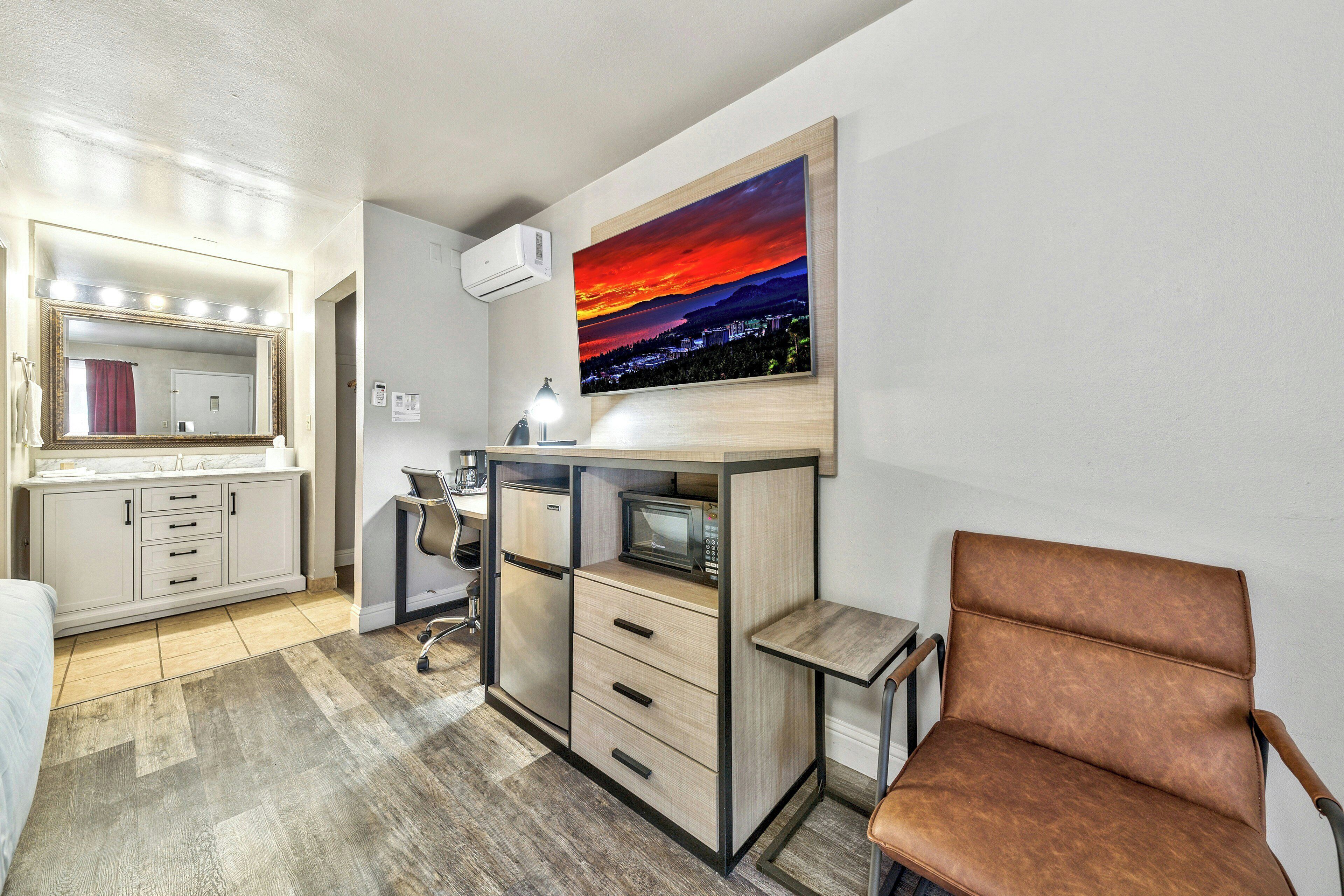 Quarto de hotel compacto com cozinha americana, um espelho grande, uma pintura de pôr do sol e uma poltrona de couro marrom.