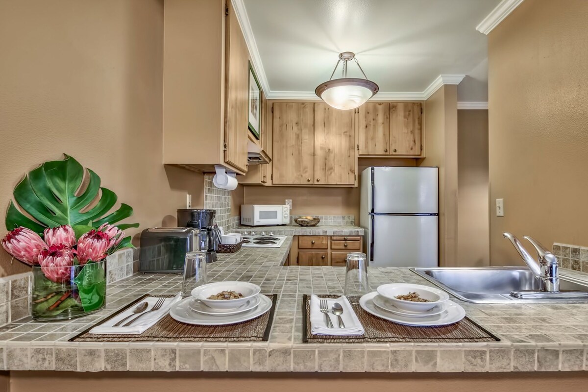 Cocina compacta con armarios de madera, un refrigerador de acero inoxidable, un microondas y una zona de comedor preparada con platos, cubiertos y un jarrón con flores.