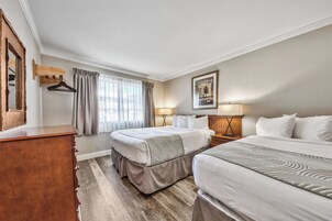 Habitación de hotel con dos camas, un aparador de madera, un espejo, una ventana con cortinas, un cuadro en la pared y una lámpara de noche.