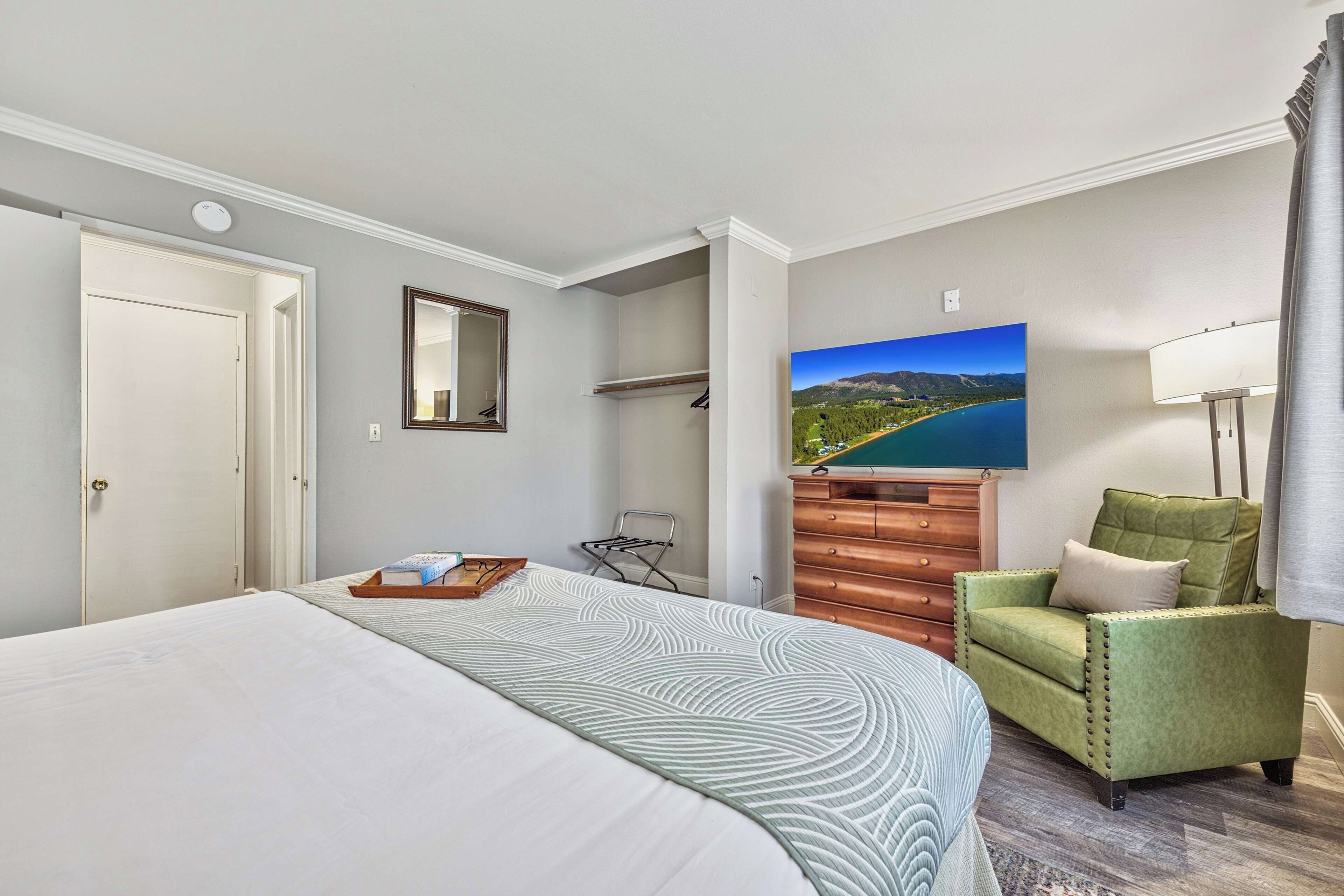A hotel room with a bed, a green armchair, a wooden dresser, a painting of a landscape, and a lamp.