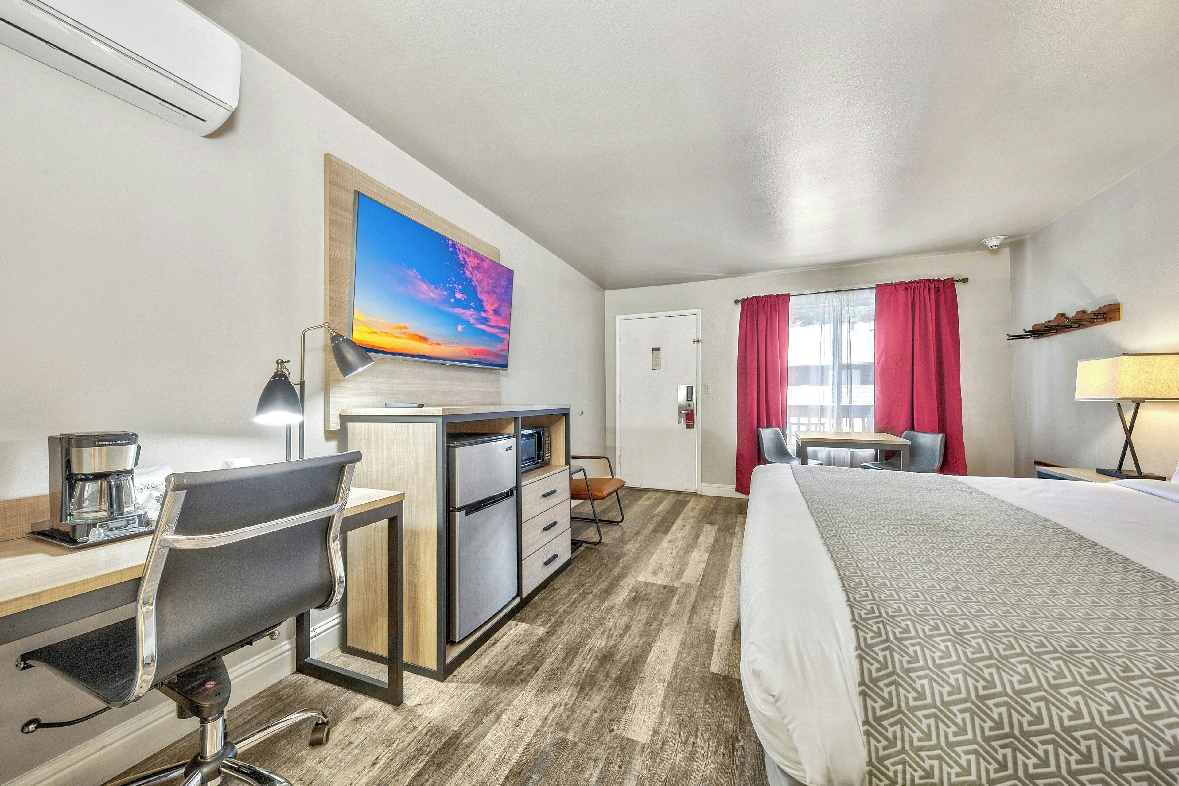 Ein Hotelzimmer mit Bett, Schreibtisch, Stuhl, Küchenzeile und einem an der Wand befestigten Fernseher.