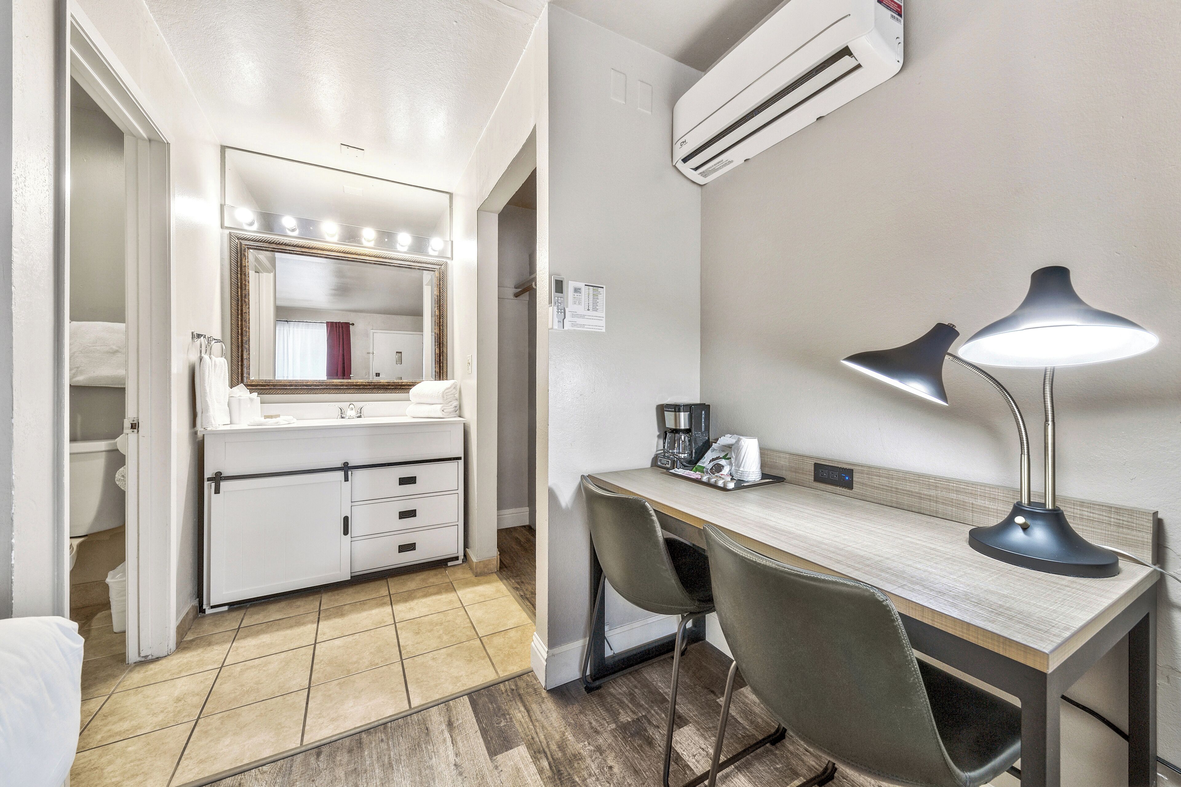 A hotel room with a bathroom, a desk with two chairs, a desk lamp, and an air conditioning unit.