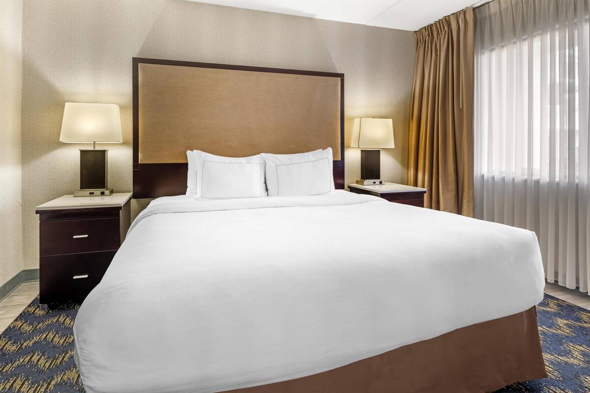 Ein Hotelzimmer mit einem großen Bett, zwei Nachttischen inklusive Lampen, einer Kopfteil und einem Fenster mit Vorhängen.