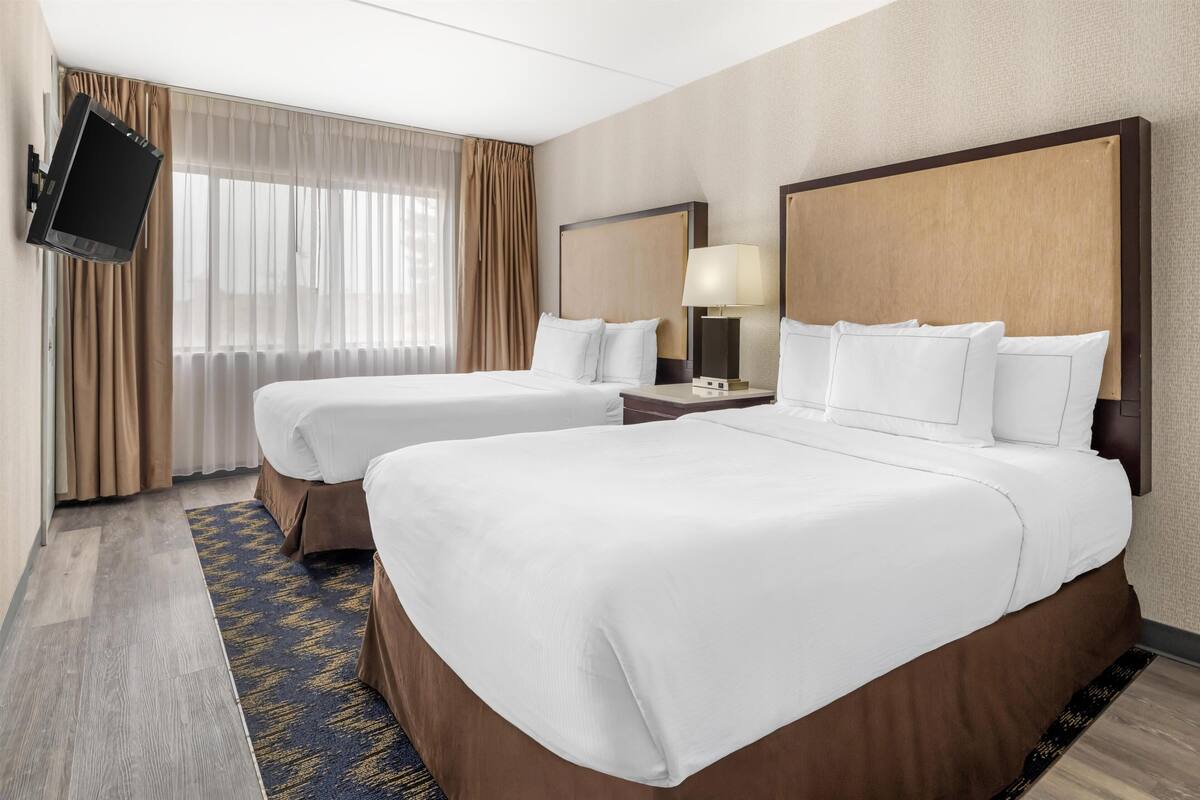 Ein Hotelzimmer mit zwei Betten, einem an der Wand befestigten Fernseher, einem Nachttisch mit Lampe und einem großen Fenster mit Vorhängen.