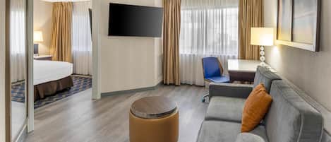 Habitación de hotel con cama, sofá, reposapiés redondo, escritorio, silla, televisor y ventana con cortinas.