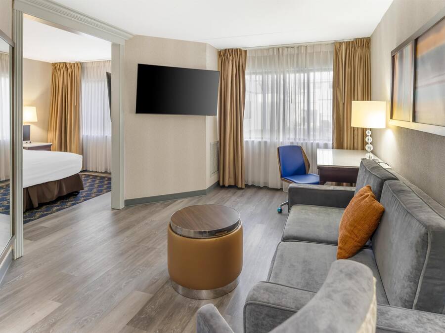 Ein Hotelzimmer mit einem Bett, einem Sofa, einem runden Hocker, einem Schreibtisch, einem Sessel, einem Fernseher und einem Fenster mit Vorhängen.