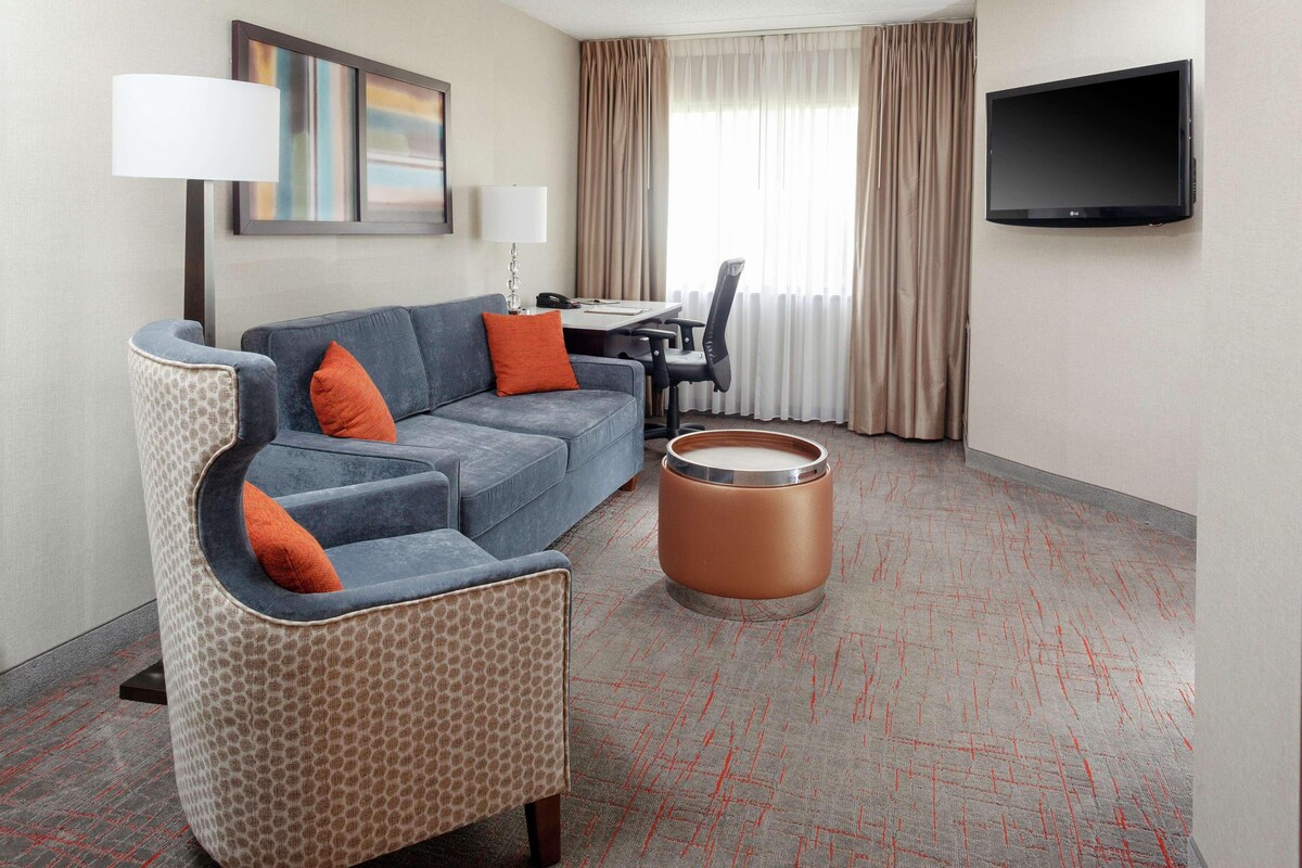 Habitación de hotel con sofá, reposapiés, mesa, silla, televisor y cortinas.