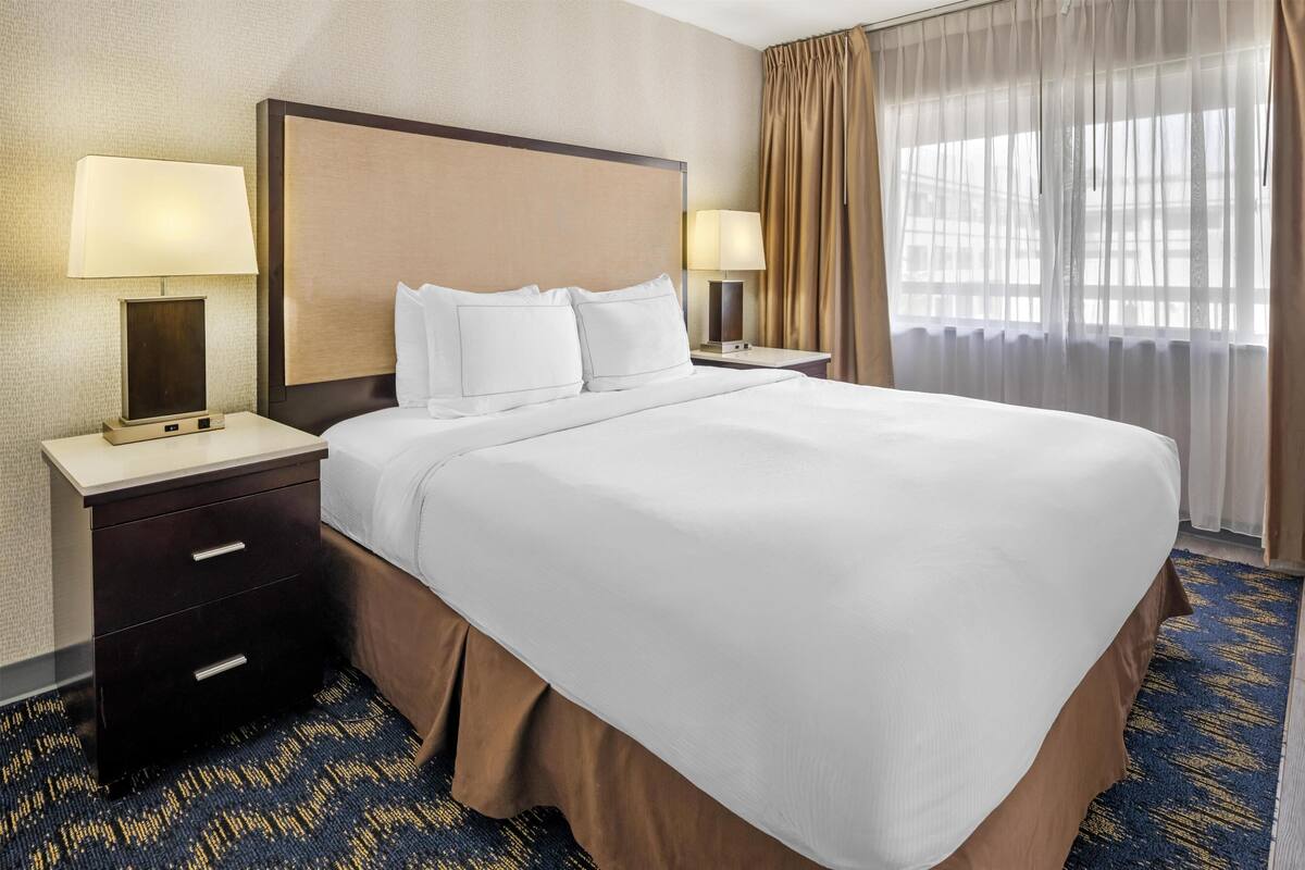 Ein Hotelzimmer mit einem großen Bett, zwei Nachttischen inklusive Lampen, einem kleinen Tischchen, einem Teppichboden und einem Fenster mit Vorhängen.